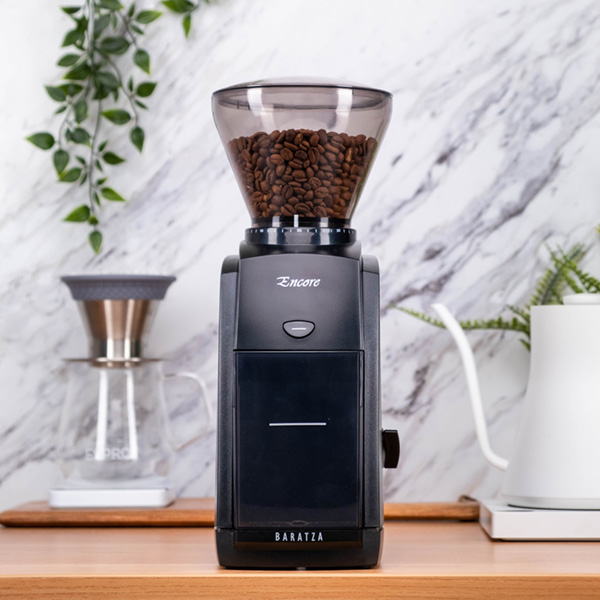 Baratza Encore Grinder sitting in front of a white marble wall with a kettle, pour over setup, and plant in the background