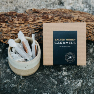 Box of San Juan Island Sea Salt Salted Honey Caramels sitting next to a cup of caramels on a piece of driftwood on a grey rock