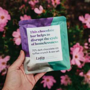Hand holding a bar of K'ul Toffee Crunch and Sea Salt Dark Chocolate over a bush with pink flowers