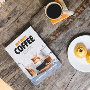 Overhead view of "But First, Coffee" book sitting on a wood table with a cup of black coffee and a pastry