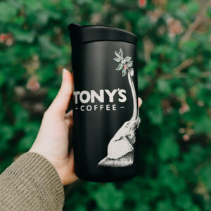Hand holding a black Tony's x Created Co. thermos in front of a forest background.